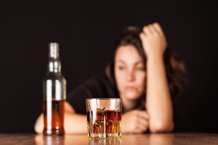 Five Dangerous Ways Alcohol Affects Your Body, upset looking young woman behind a bottle and full glass of liquor - alcohol addiction